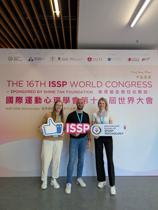 ISSP_AB Sportpsychologie Gruppenfoto: 3 Menschen stehen nebeneinander vor einer Leinwand mit dem ISSP Log und lachen in die Kamera