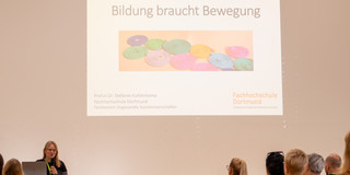 Das Bild zeigt hauptsächlich die Titelfolie einer Präsentation mit dem Titel Bildung braucht Bewegung. Außerdem sind die Rednerin und ein kleiner Teil der Besucher*innen im unteren Teil des Bildes zu sehen.