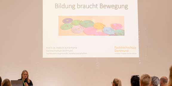 Das Bild zeigt hauptsächlich die Titelfolie einer Präsentation mit dem Titel Bildung braucht Bewegung. Außerdem sind die Rednerin und ein kleiner Teil der Besucher*innen im unteren Teil des Bildes zu sehen.