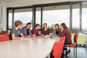 Six students are sitting at a table with books and notepads on.